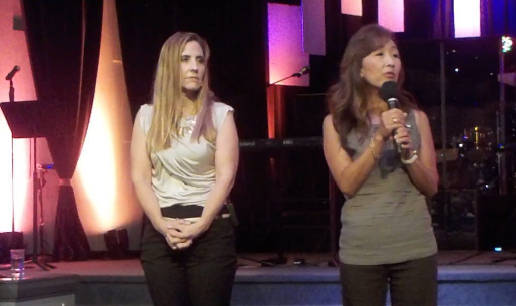 Two ladies with sincere expressions, one lady is speaking into a handheld microphone