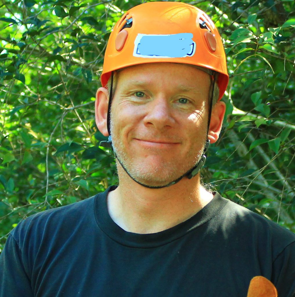 A man standing with a hard hat used for mountain climbing, with a three-day growth of beard and calm smile