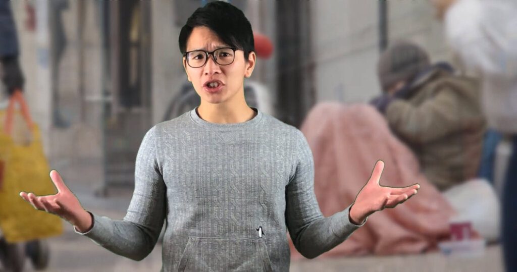 Young man with glasses wearing a long-sleeve shirt standing in front of a picture of homeless people, gesturing with both hands and speaking.